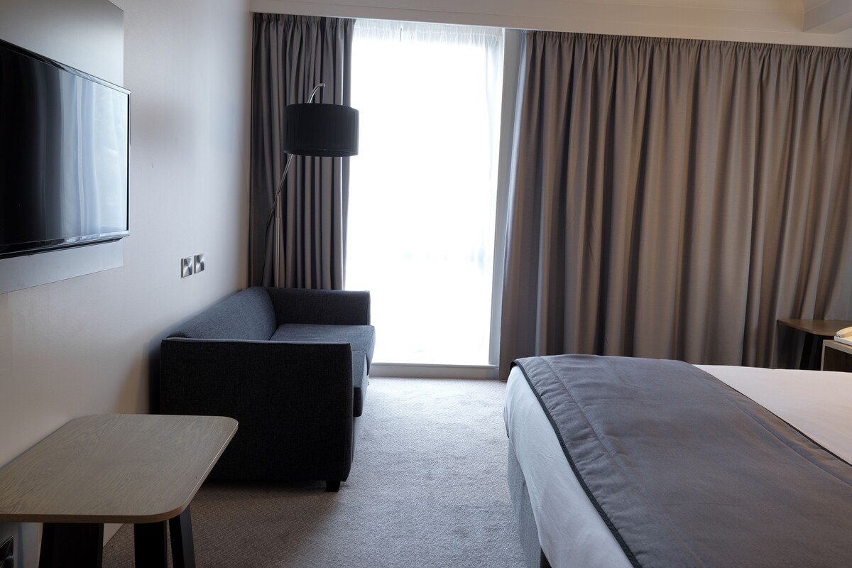 Habitación de hotel con cama, sofá, televisor y ventana con cortinas.