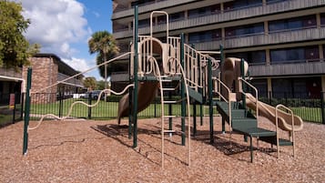 A playground with a slide and climbing structures, surrounded by a brick building and a fence.