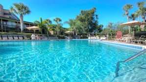 A clear blue pool with lounge chairs and umbrellas, surrounded by palm trees and a building with a balcony.
