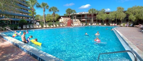 A swimming pool with people in it, surrounded by palm trees and a building with a sign that reads "Hotel".