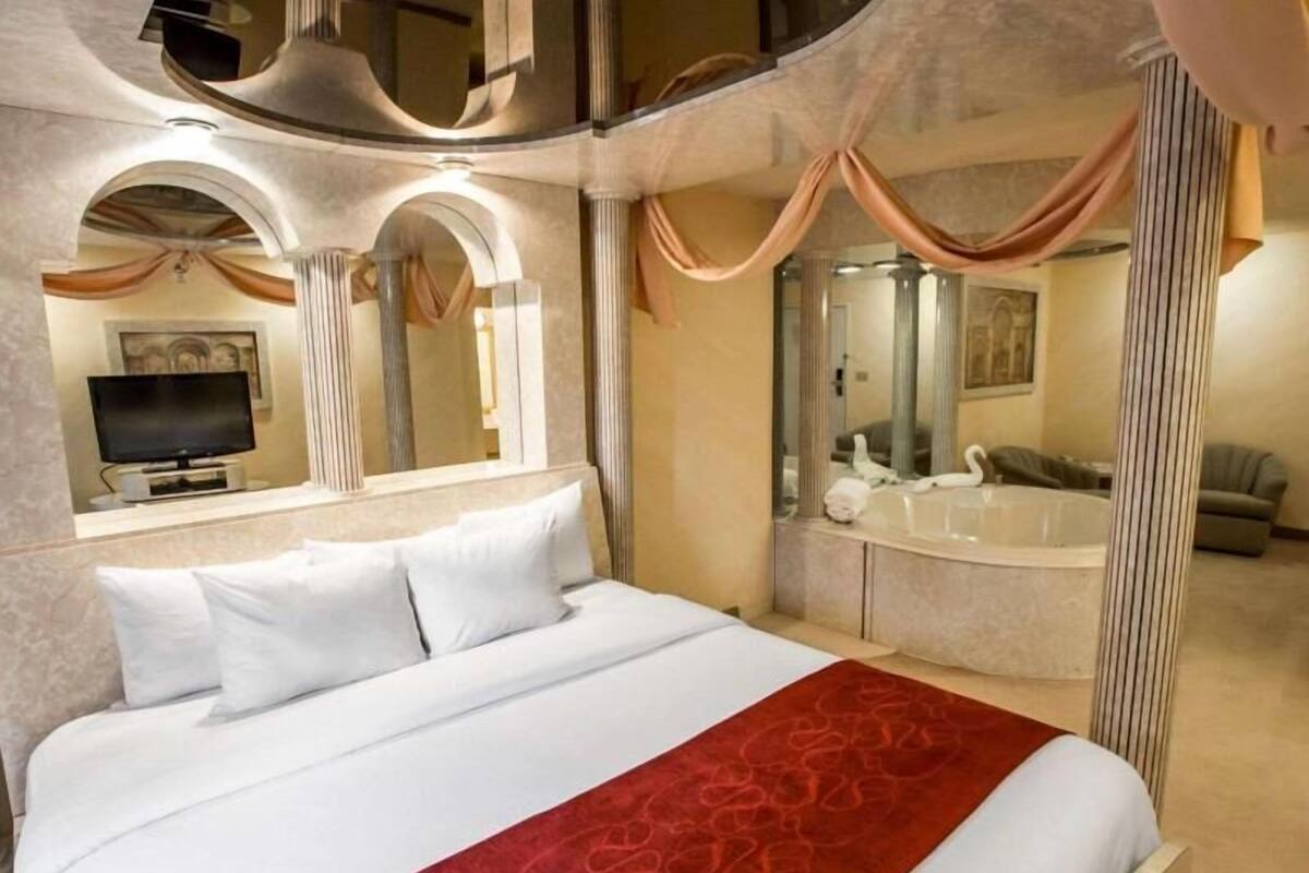 A hotel room with a large bed, a TV, a bathtub, and a mirror.