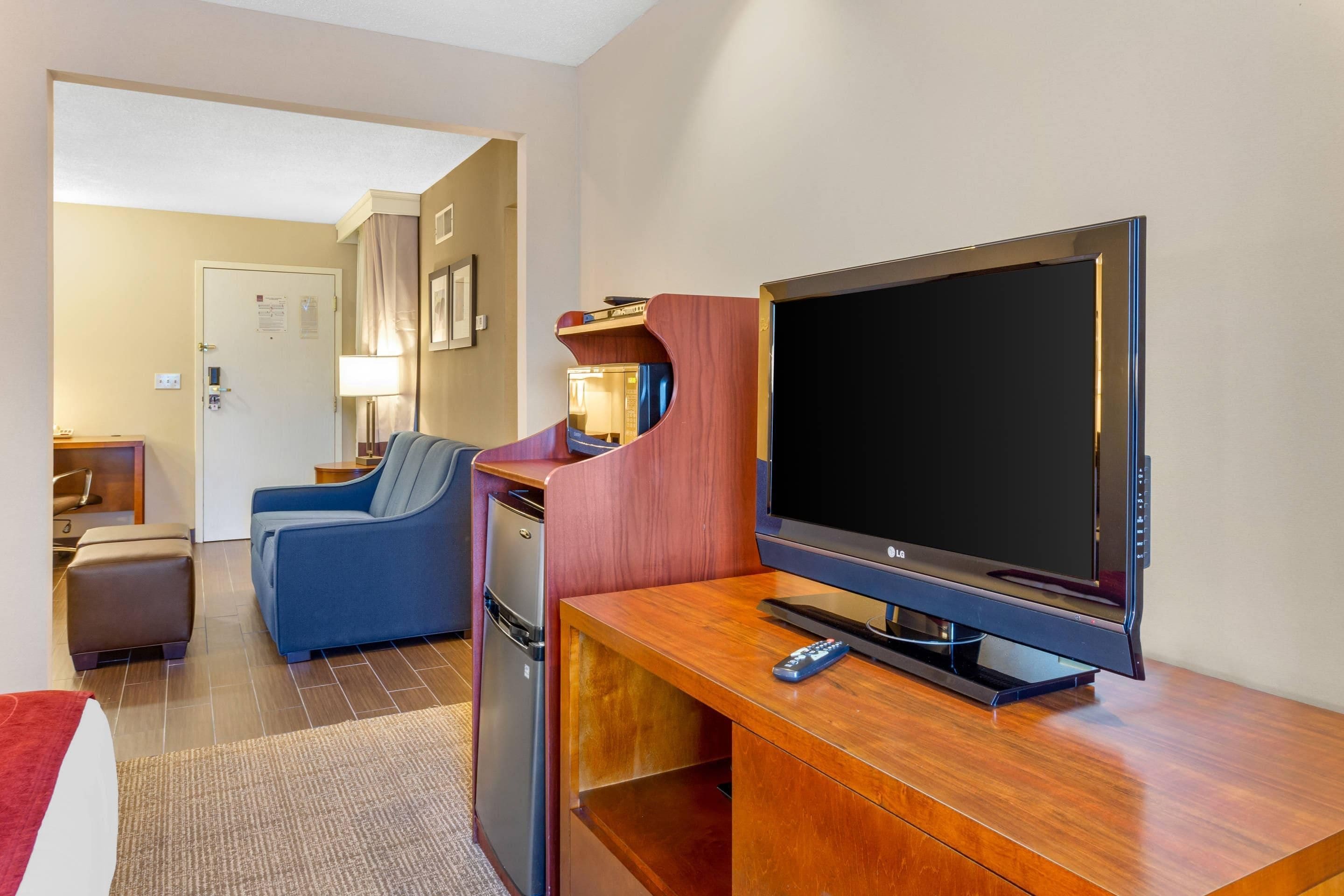 A hotel room with a flat-screen TV, a sofa, a small table, and a refrigerator.