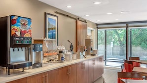 A modern kitchen area with a coffee machine, a juice dispenser, and a large window providing a view of the outdoors. - Comfort Suites Oakbrook Terrace - Chicago West (Villa Park)