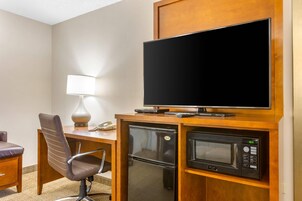 Habitación de hotel con un escritorio de madera, televisor, microondas, refrigerador, silla y mesita de noche.