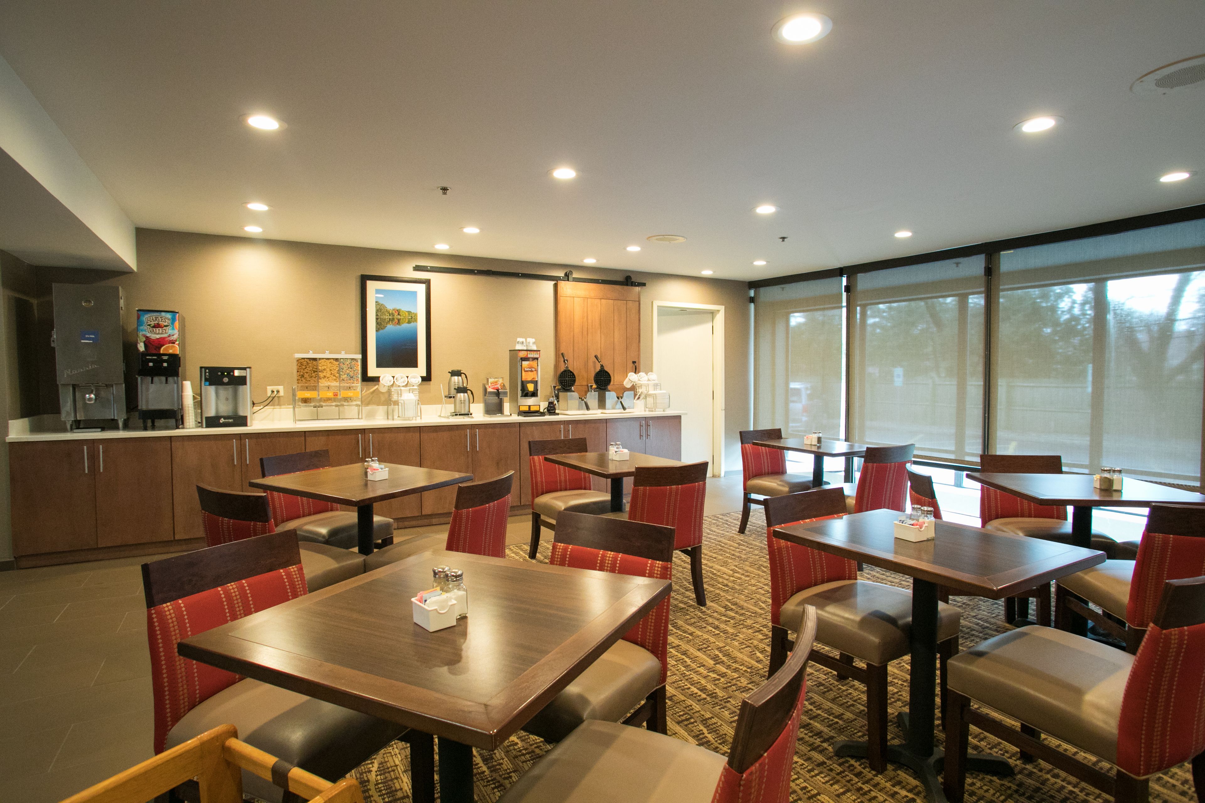Espaço de refeições de hotel com várias mesas e cadeiras, área de cozinha com eletrodomésticos e grandes janelas que proporcionam luz natural.