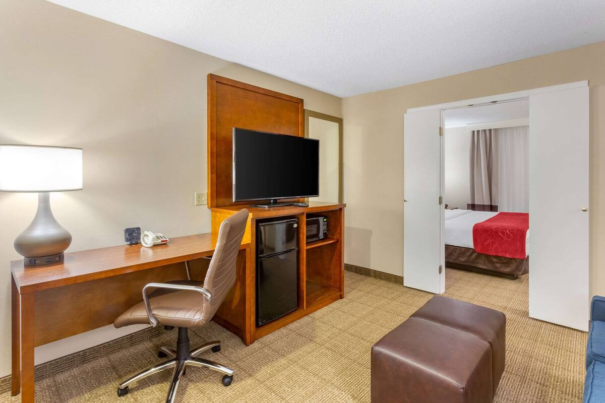 A hotel room with a wooden desk, a television, a refrigerator, a chair, and a bed with a red blanket.