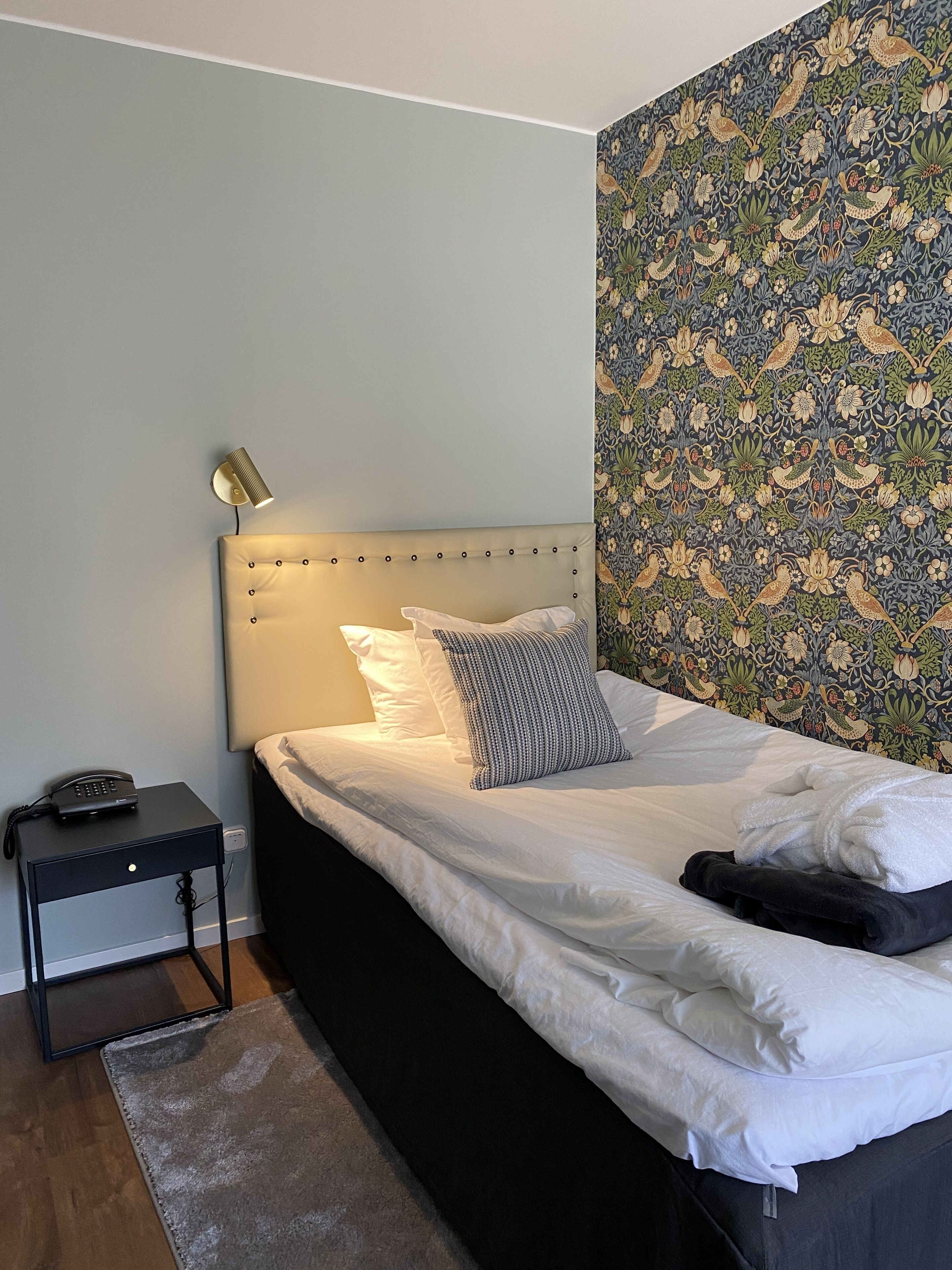 Een net opgemaakt bed met witte lakens en een gestreept kussen, een bedlampje, een zwart nachtkastje met een telefoon, en behang met een patroon.