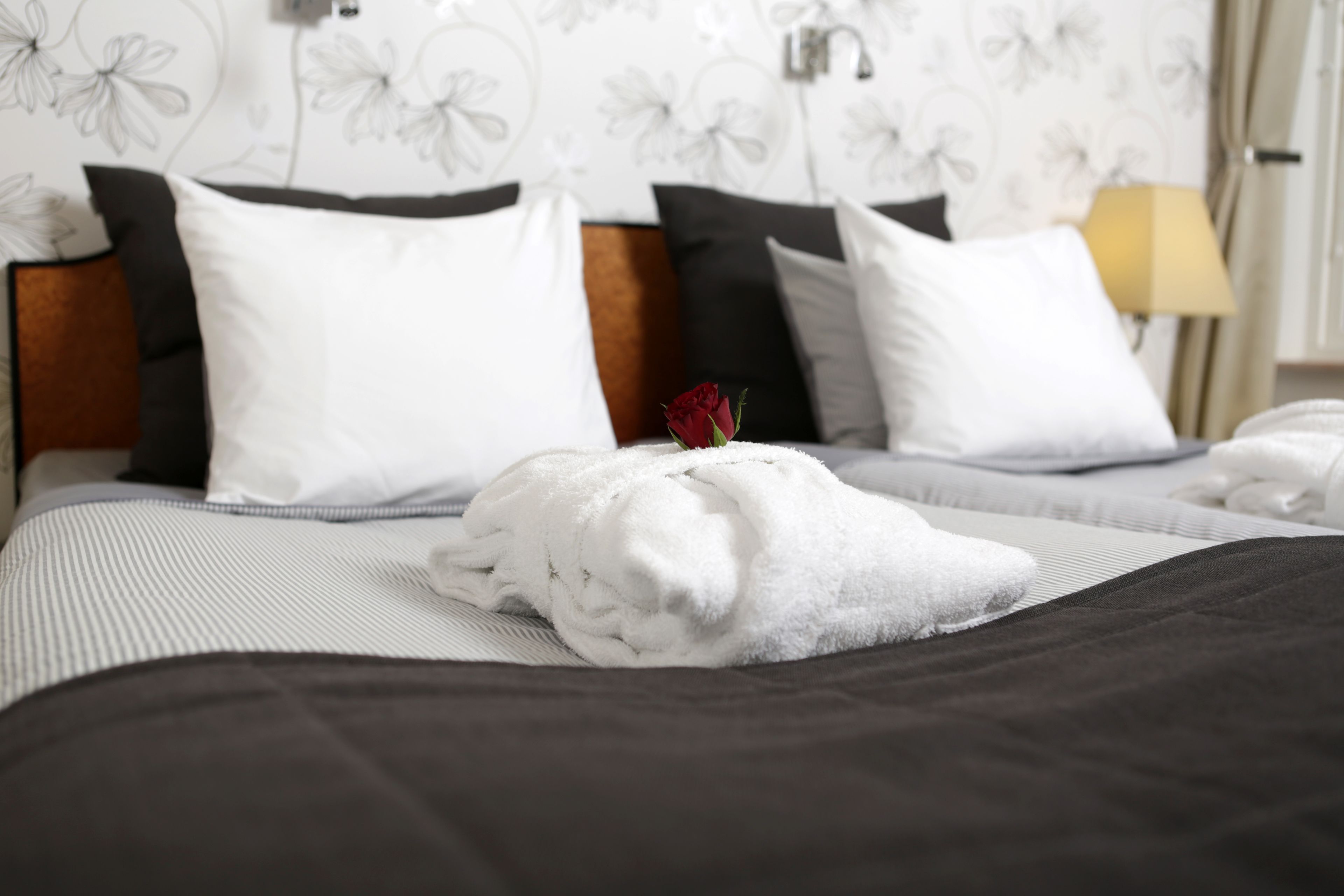 Una cama bien hecha con una toalla blanca y una rosa roja, almohadas negras y blancas, y un cabecero de madera.