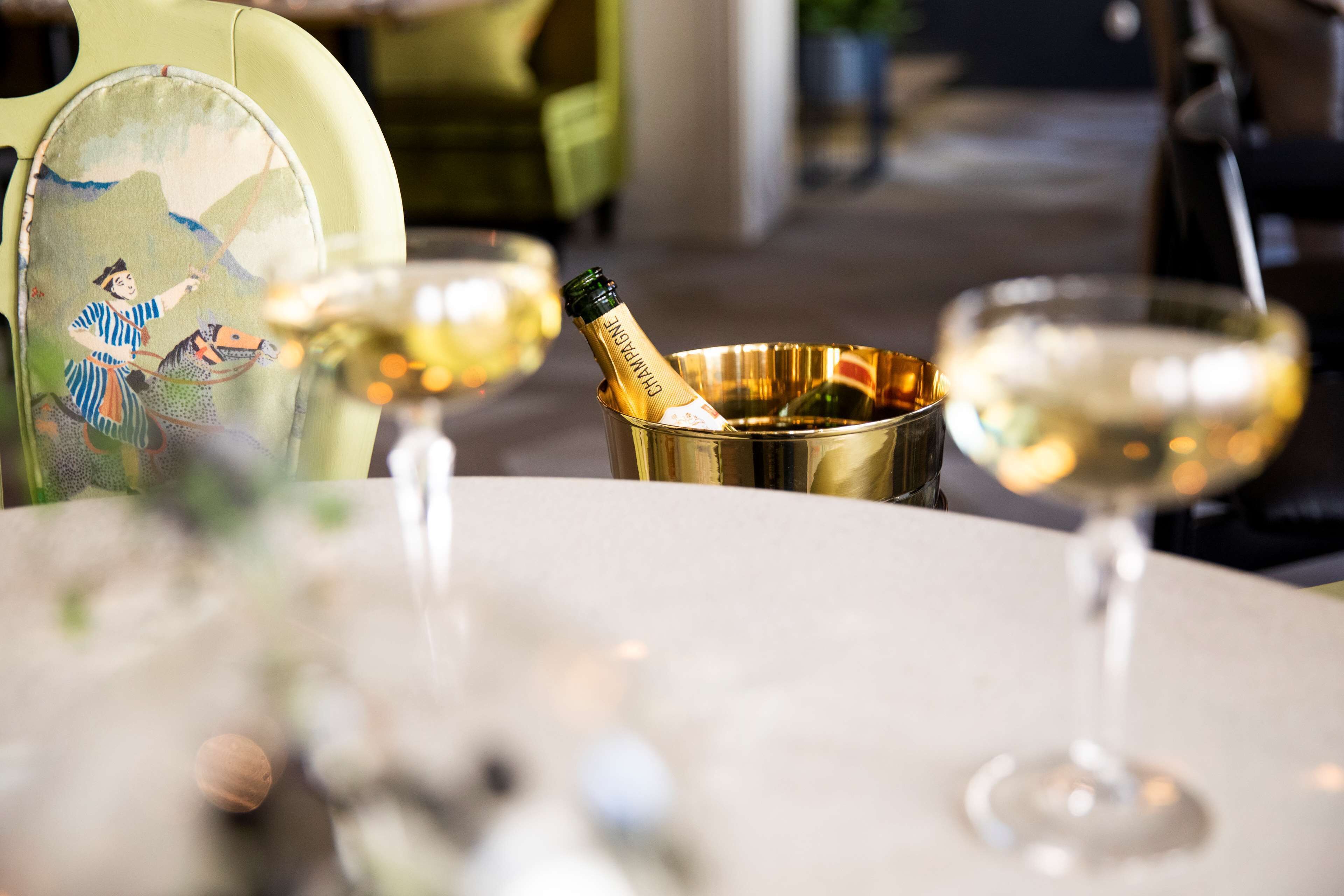 A table with a bottle of champagne in a gold bucket, a glass of champagne, and a decorative chair with a traditional design.