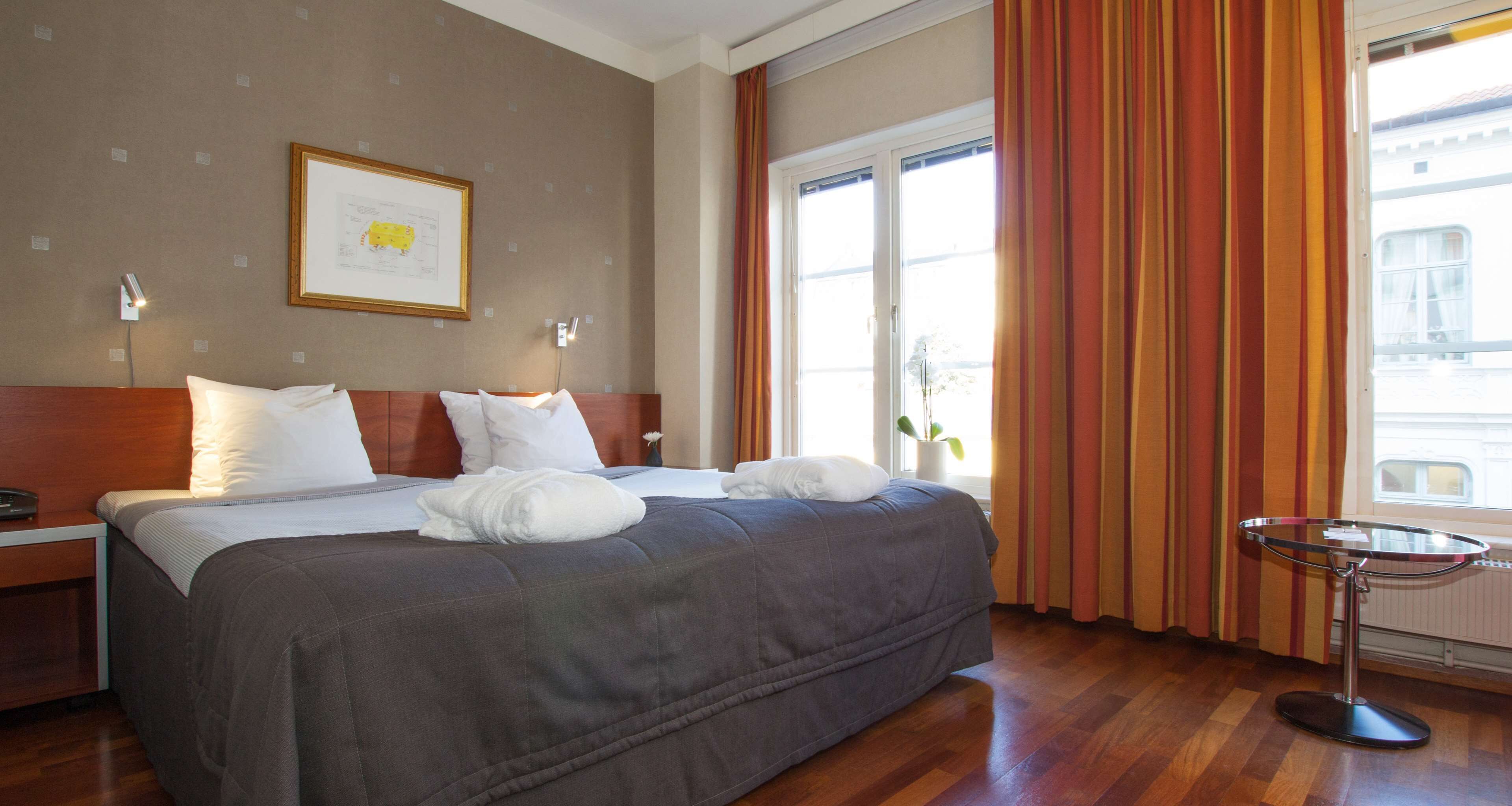 Una habitación de hotel con una cama grande, una mesita de noche, una mesa redonda pequeña y una ventana con cortinas.