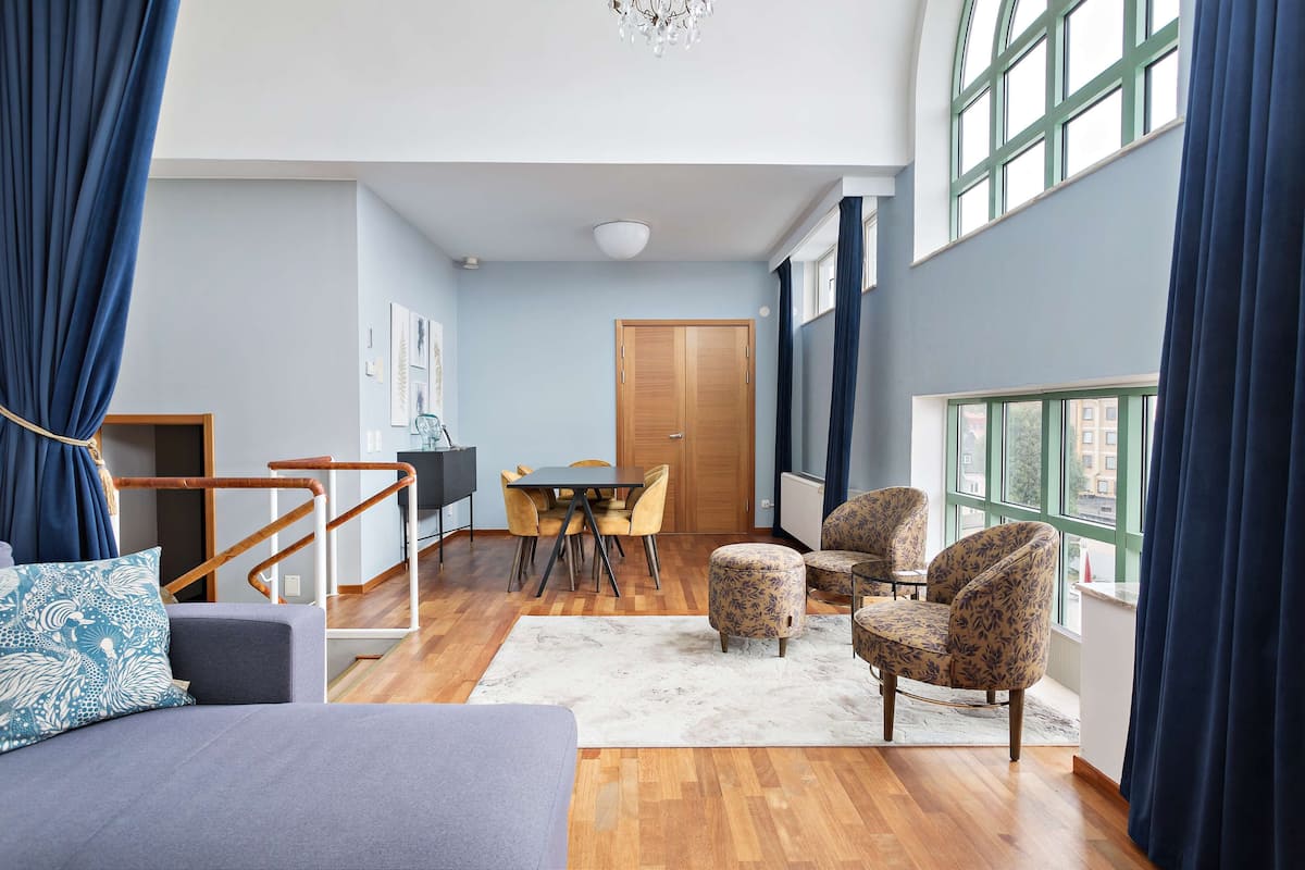 A spacious living area with a sofa, ottoman, and chairs, a dining table with chairs, a staircase, and large windows with curtains.