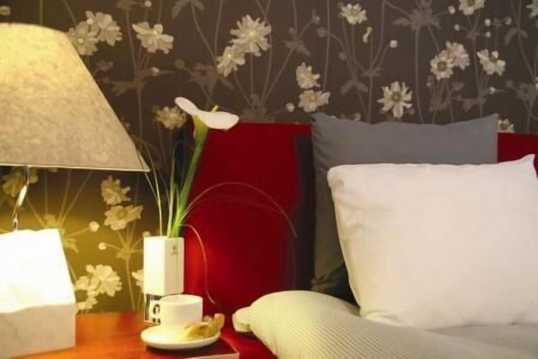 A hotel room with a red headboard, a white pillow, a bedside table with a lamp, a vase with flowers, and a cup and saucer.