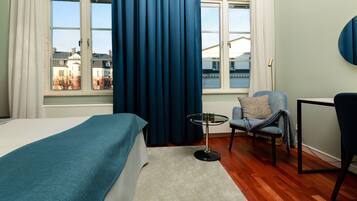 A hotel room with a bed, a small round table, a chair, and a view of buildings through large windows.