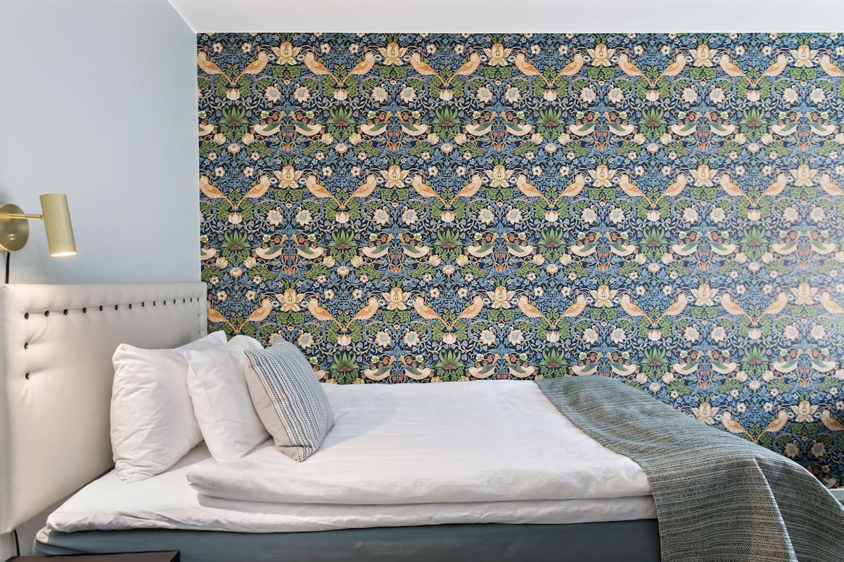 A bedroom with a patterned wallpaper, a tufted headboard, white bedding, and a bedside lamp.