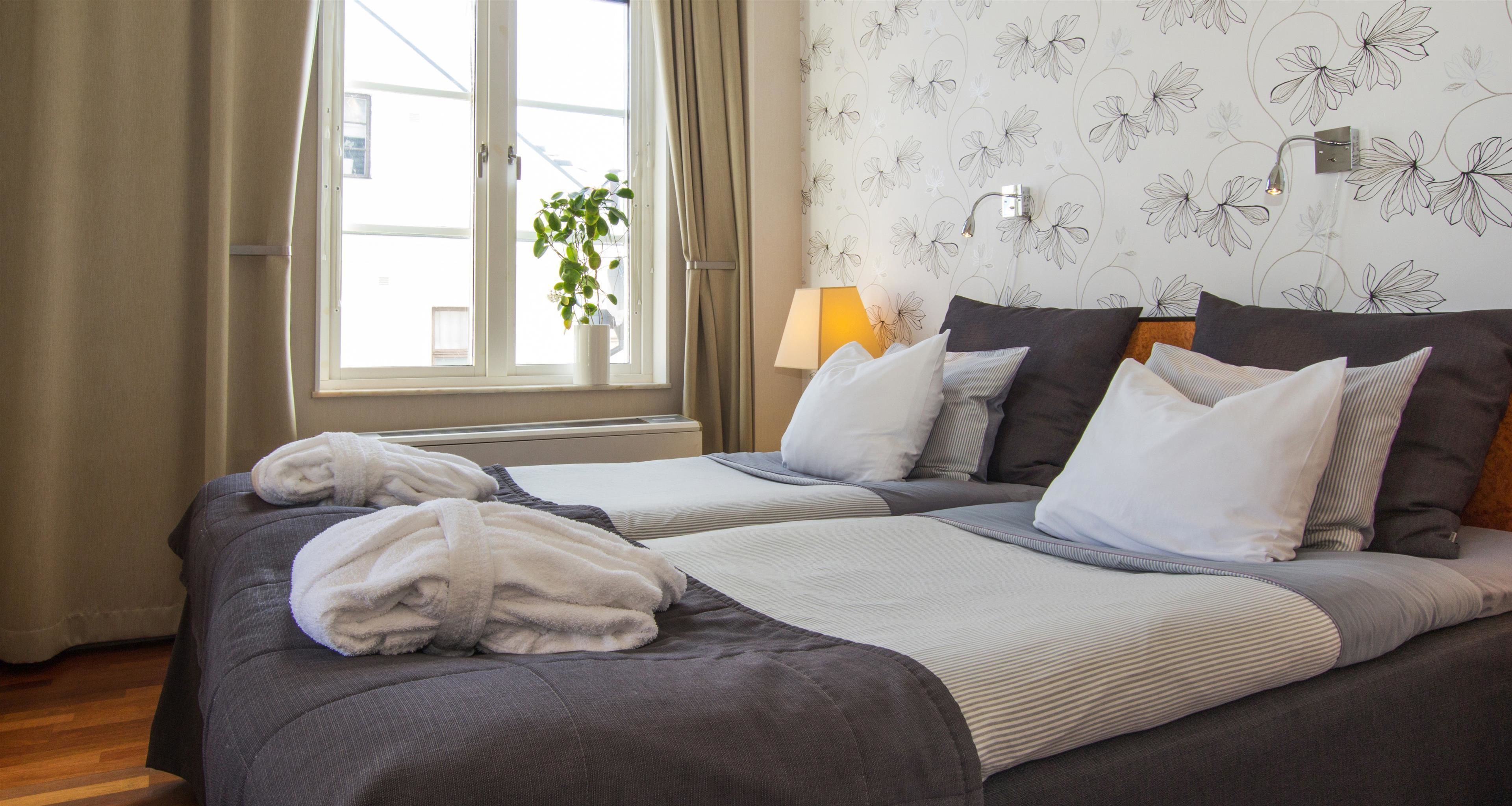 Ett hotellrum med två sängar, ett fönster med gardiner, en växt och en vägg med ett blommönster.