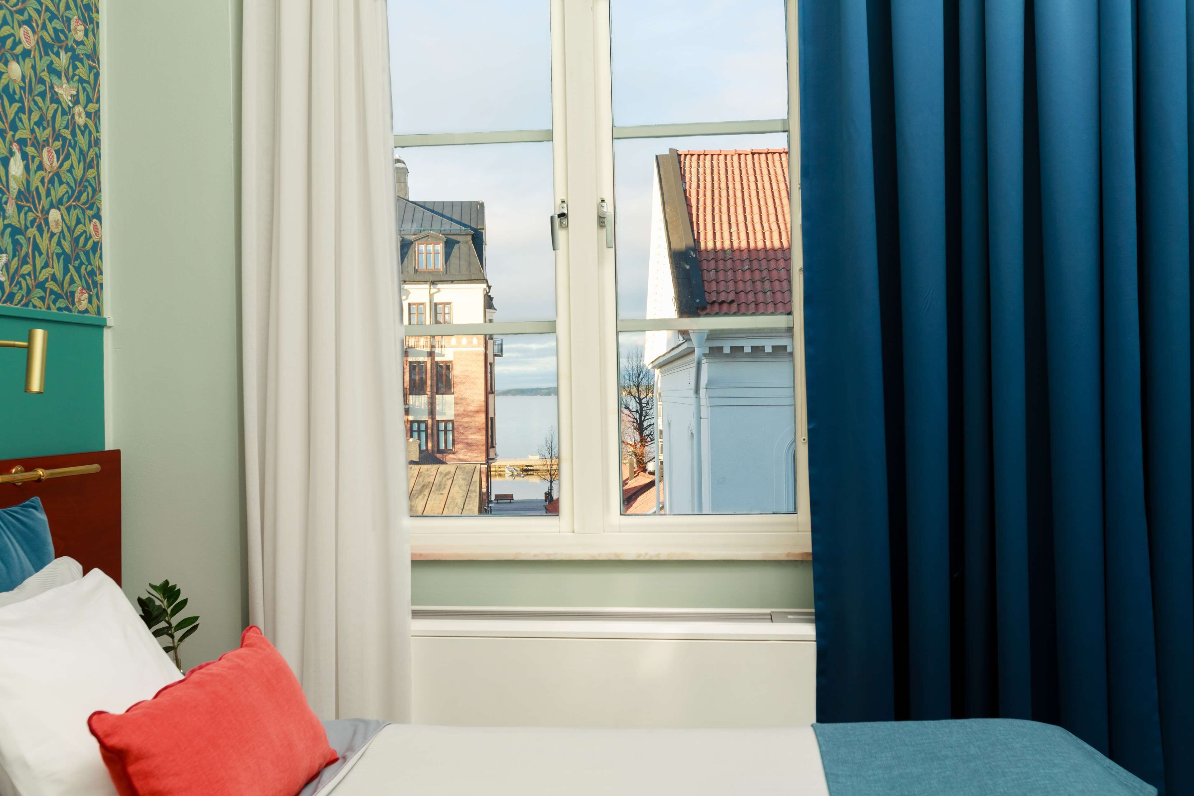 A hotel room with a large window, a bed with white and red pillows, and a view of buildings outside.
