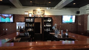 A bar area with a wooden counter, multiple shelves of liquor bottles, and two TVs mounted on the walls.