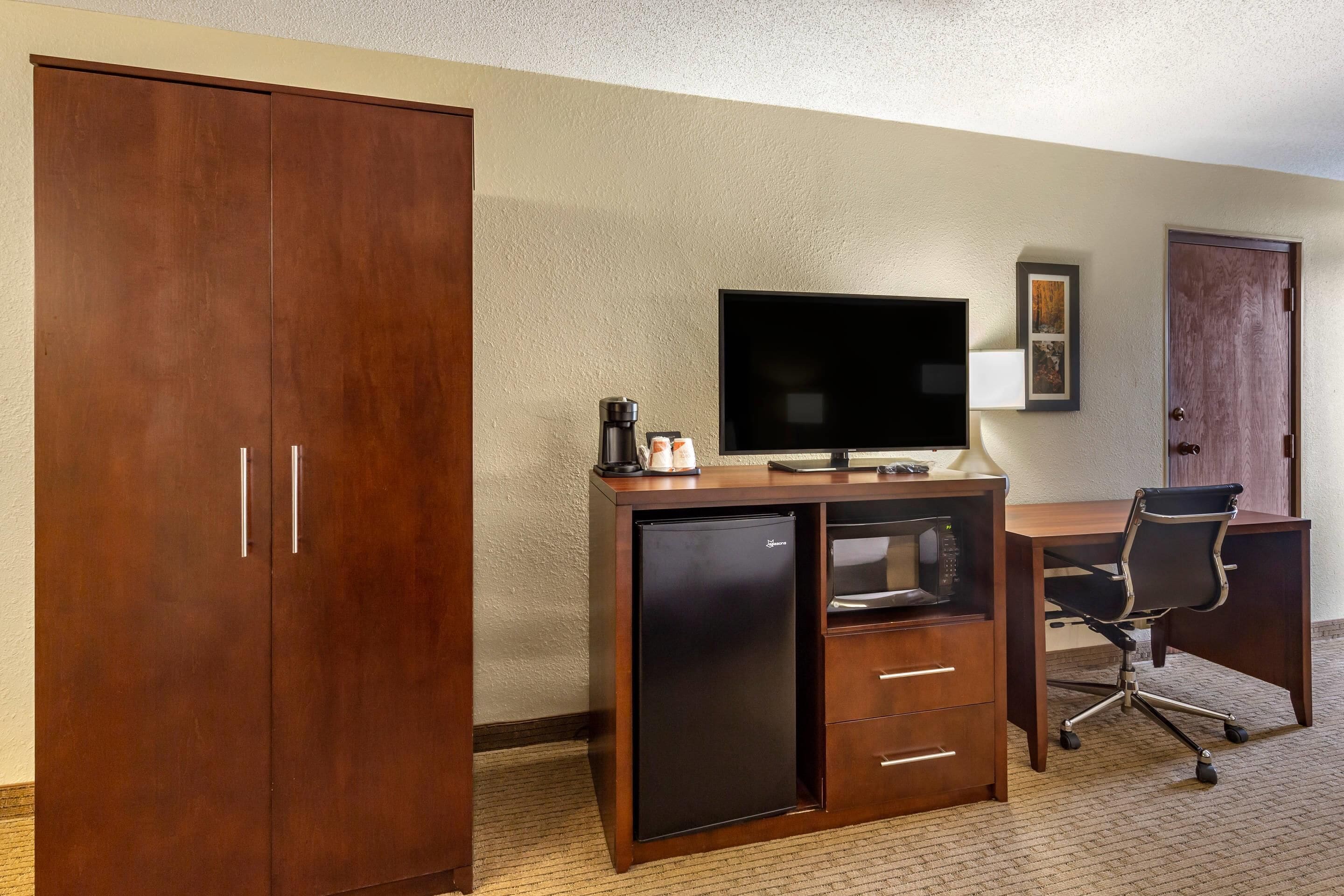 Ein Hotelzimmer mit Holzschrank, Schreibtisch mit Computer, Stuhl, Fernseher und einer Kaffeemaschine.