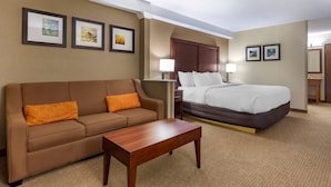 A hotel room with a brown sofa, a wooden coffee table, a bed with white bedding, and framed artwork on the walls.