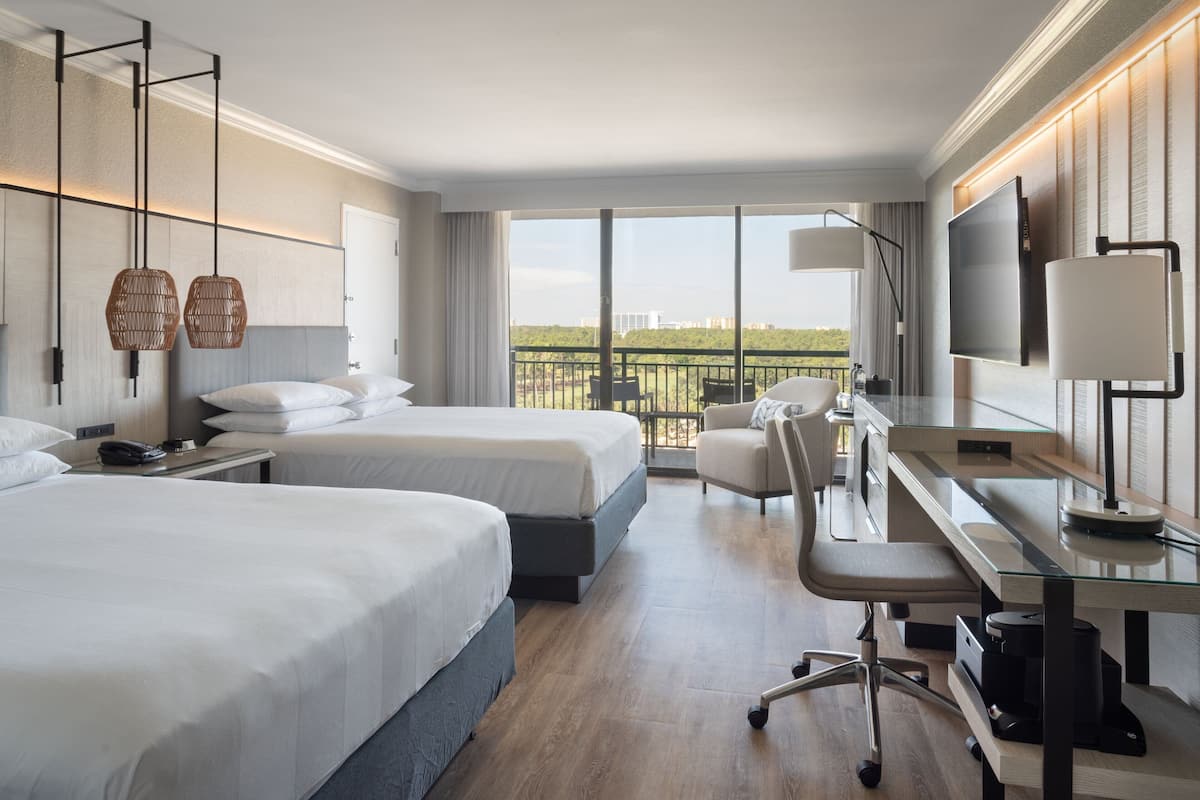 A hotel room with two beds, a desk, a chair, a TV, and a balcony with a view.
