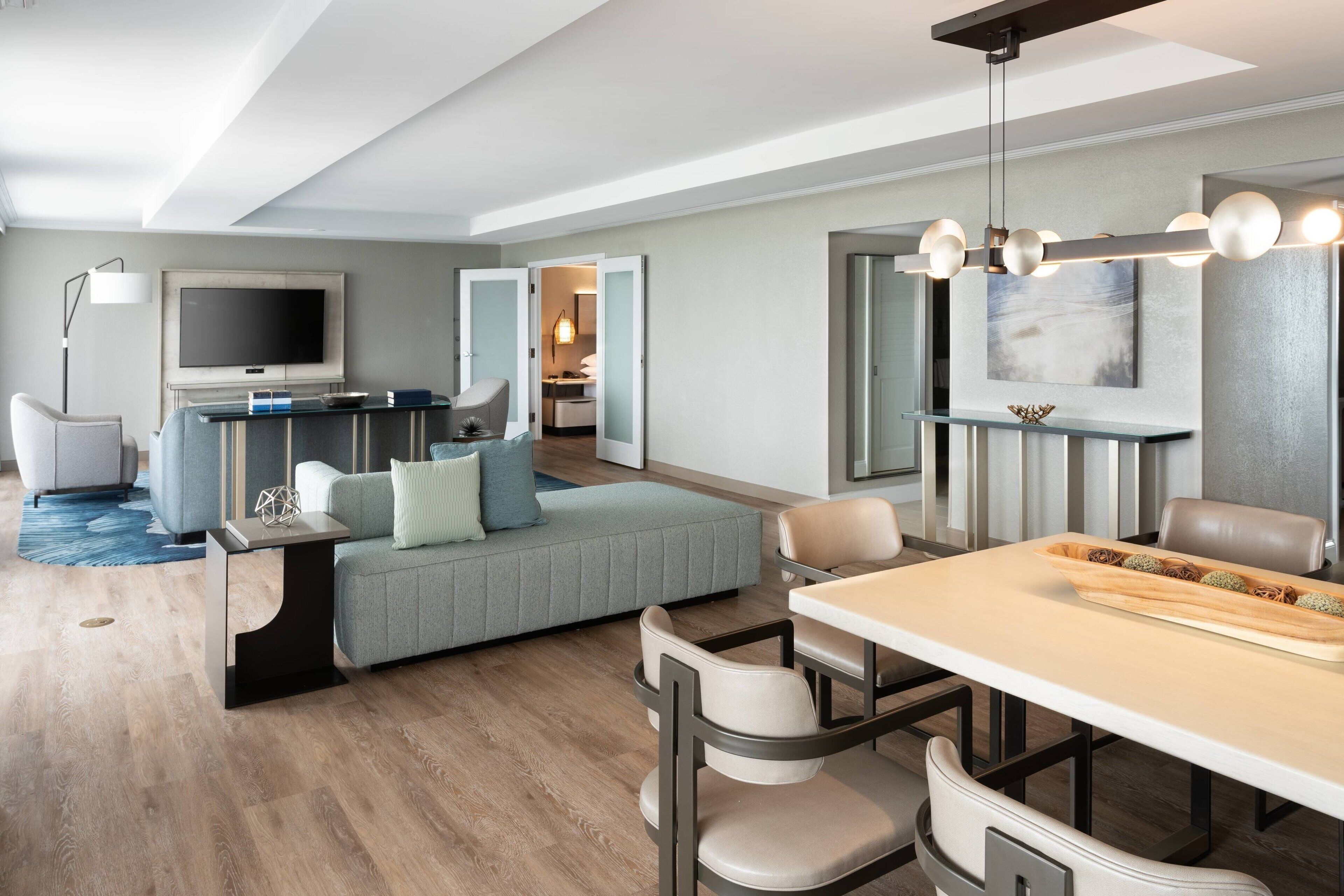 A modern hotel room with a sofa, dining table, chairs, a TV, and a large mirror.