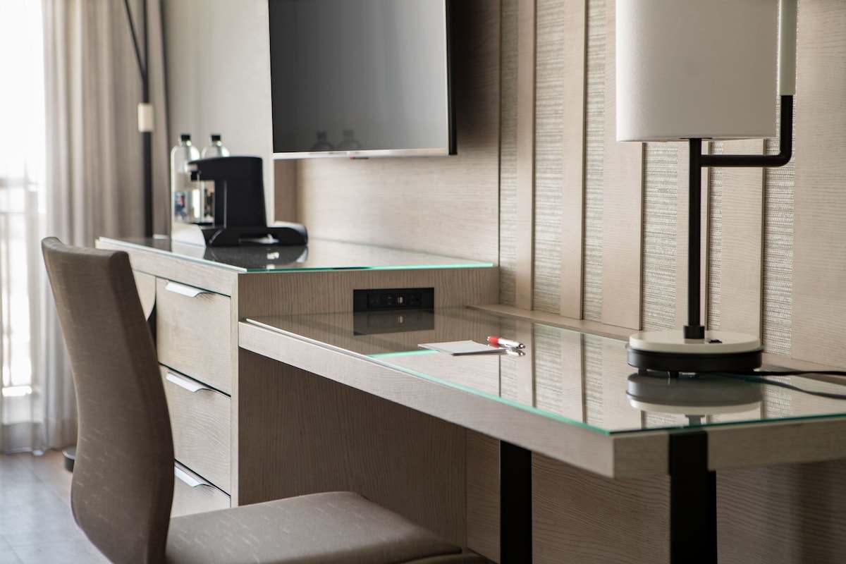 A modern hotel room with a glass desk, a flat-screen TV mounted on the wall, and a minimalist lamp.