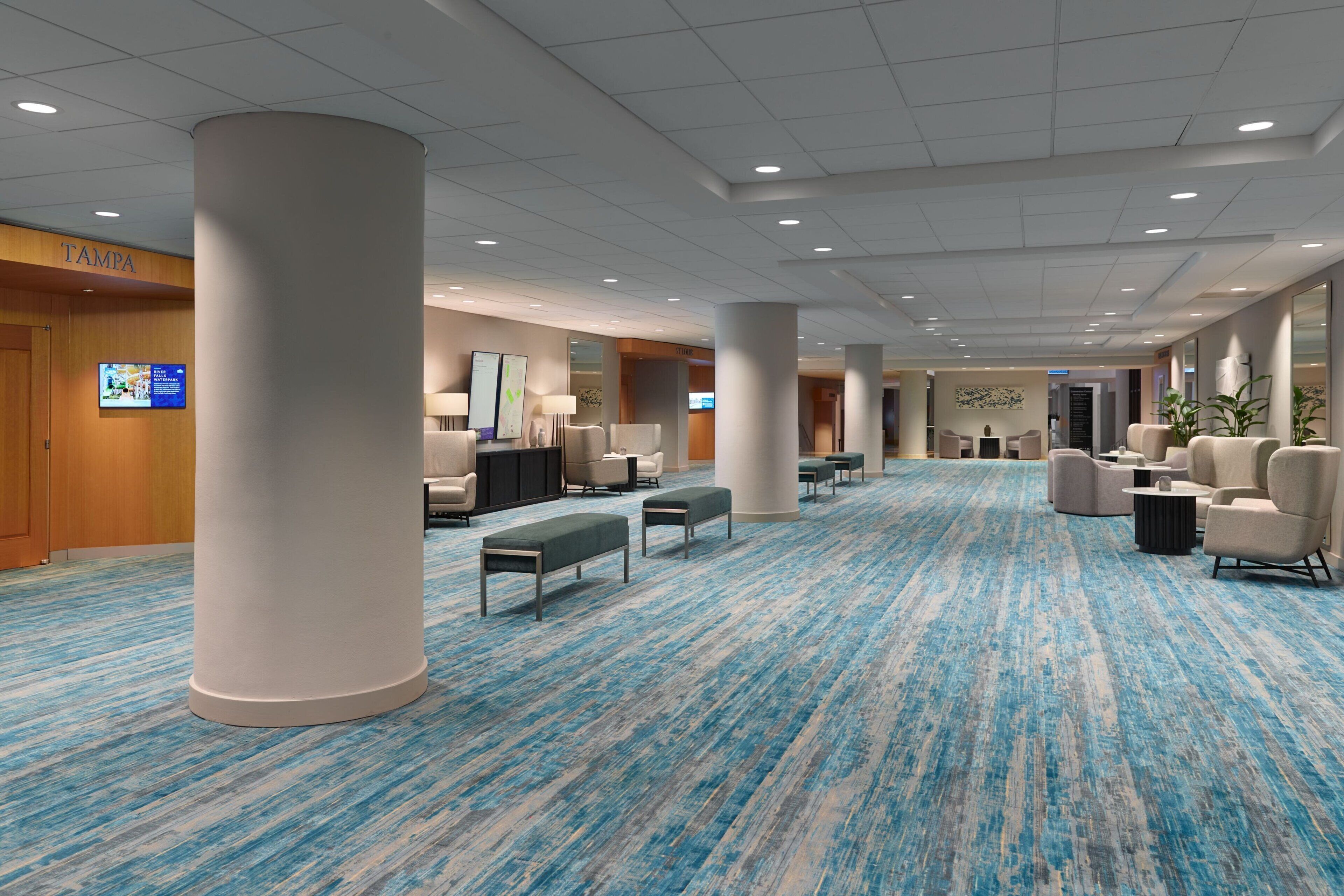 Een ruime hotel lobby met een blauw tapijt, verschillende zithoeken en een grote pilaar.