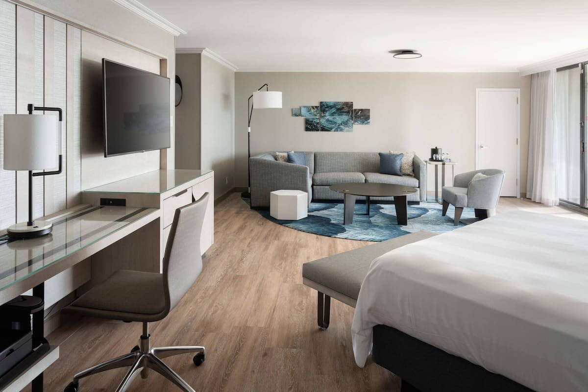 A modern hotel room with a flat-screen TV, a glass desk, a grey sofa, a round coffee table, and a bed with white bedding.