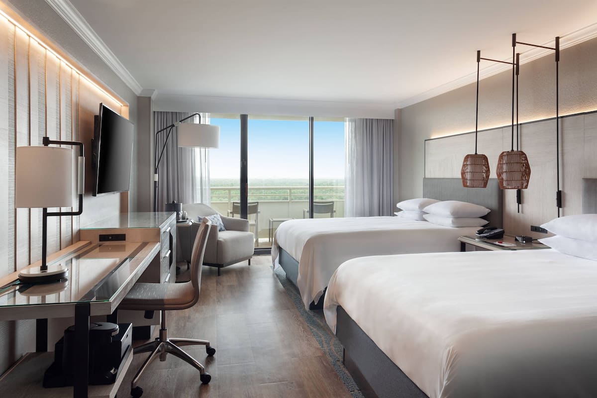 A hotel room with a desk, two beds, a chair, a TV, and a large window with a view.