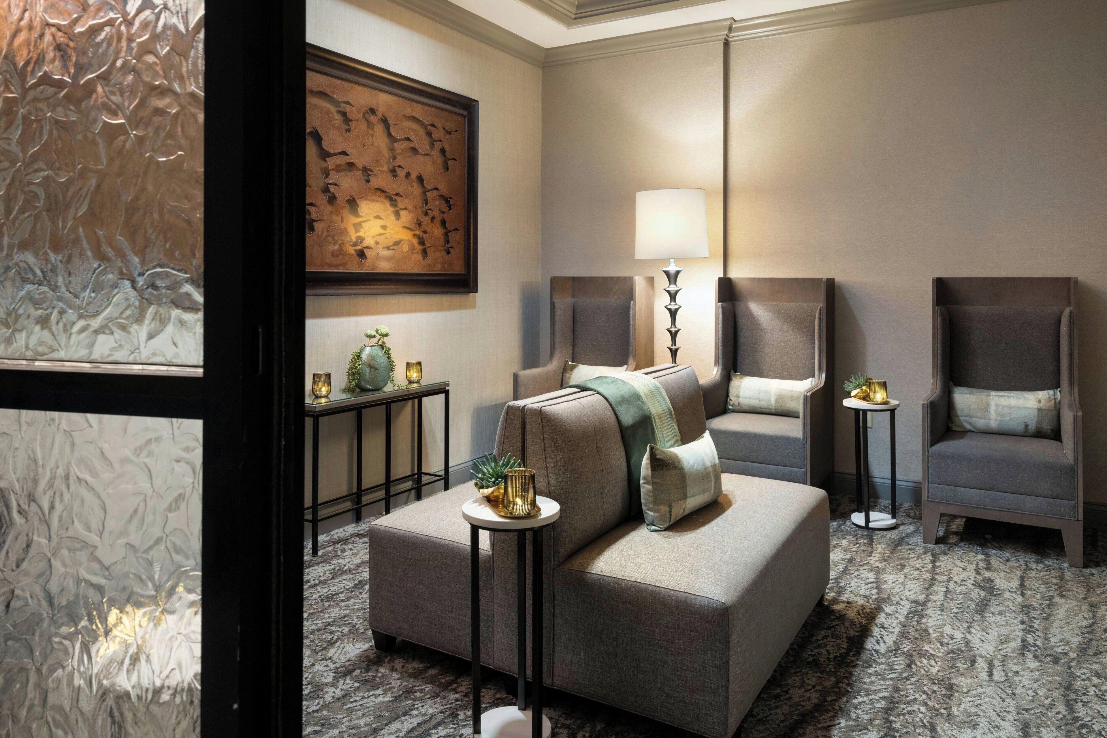 A modern hotel room with a sofa, armchairs, a lamp, a painting, and a small table with a plant.