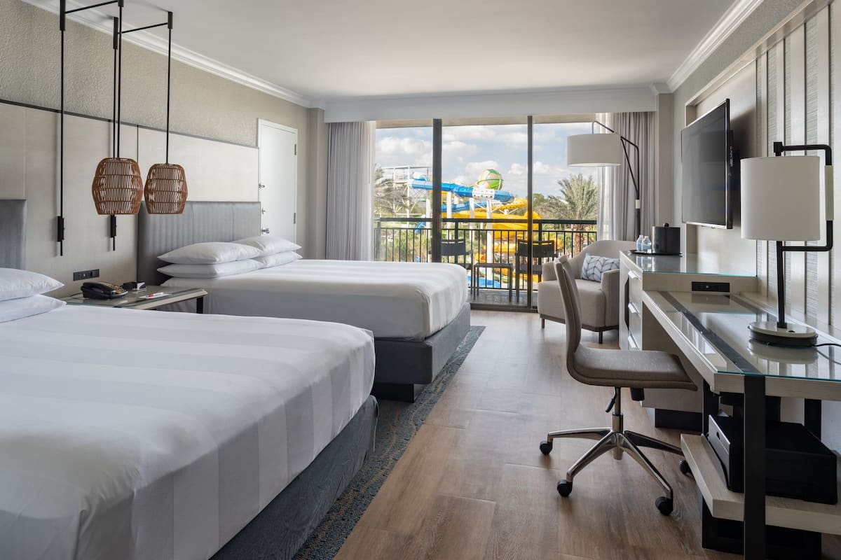 A hotel room with two beds, a desk with a chair, a TV, and a balcony with a view of a water park.