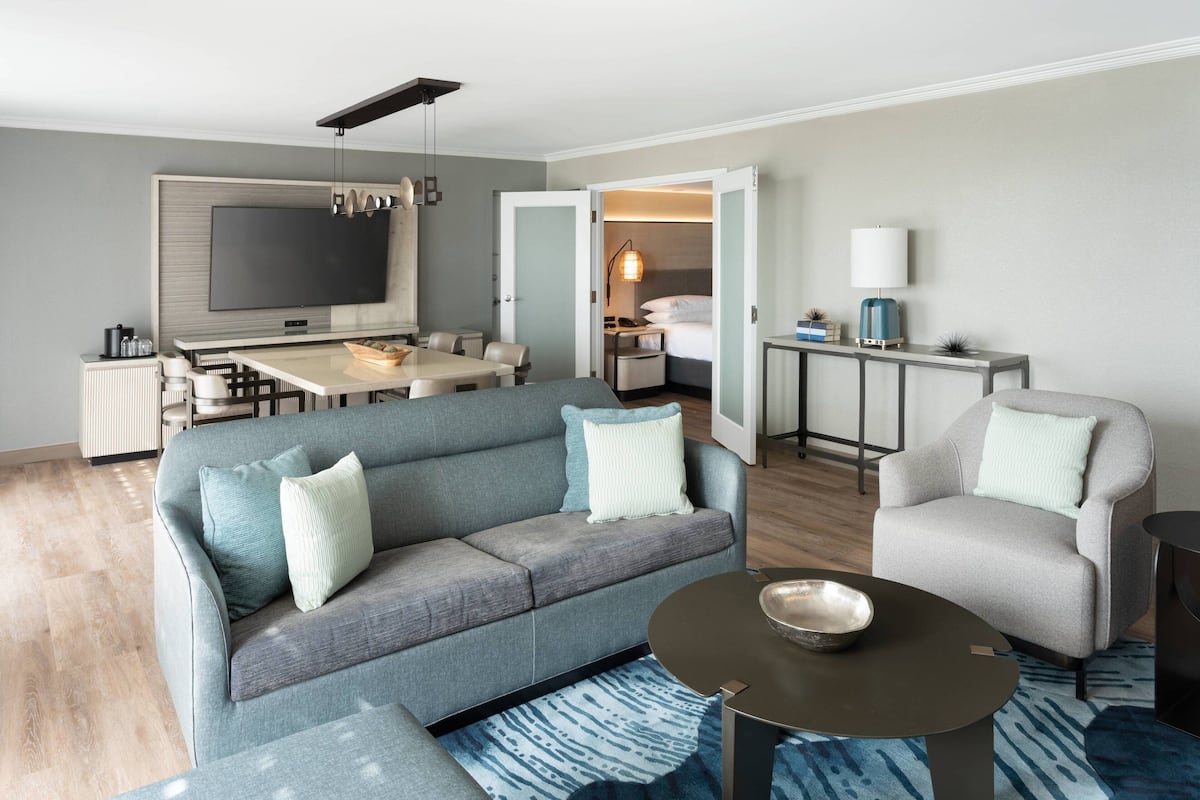 A modern hotel room with a grey sofa, a round coffee table, a dining area with a TV, and a separate bedroom with a bed and bedside table.