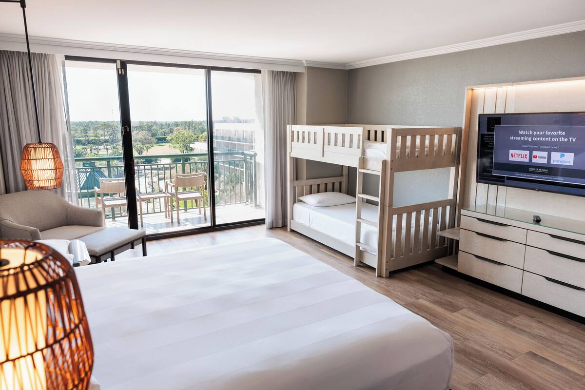 A hotel room with a large window, a bed, a TV, a sofa, and a balcony.