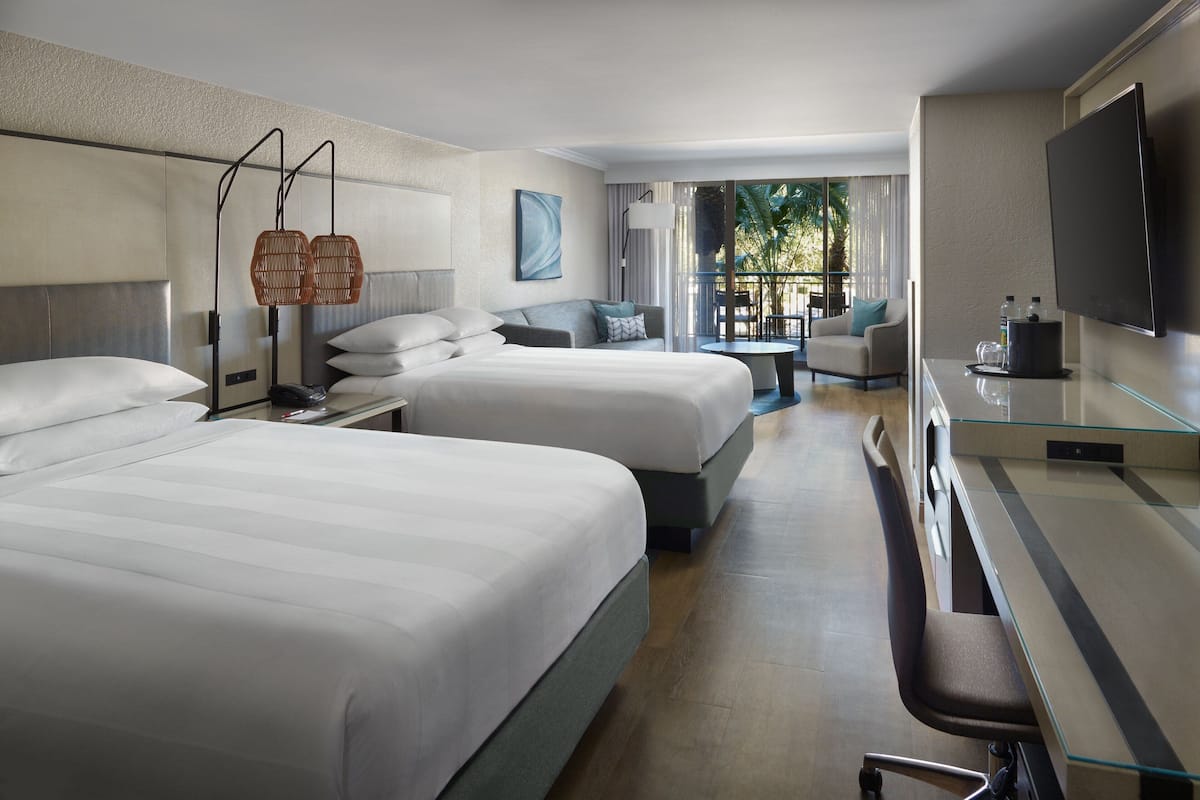 A hotel room with two beds, a desk, a chair, a TV, and a balcony with a view of palm trees.