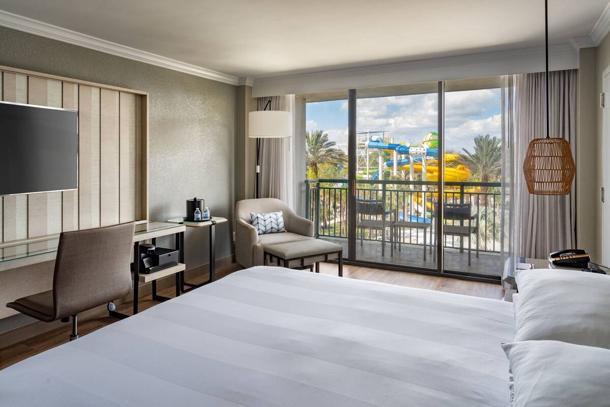 A hotel room with a large bed, a desk, a chair, a TV, and a balcony with a view of a water park.