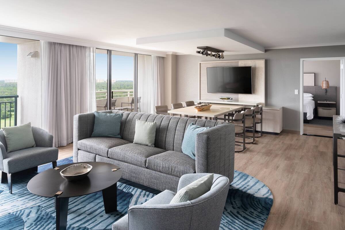 A modern hotel room with a large sofa, a dining table, a TV, and a balcony with curtains.