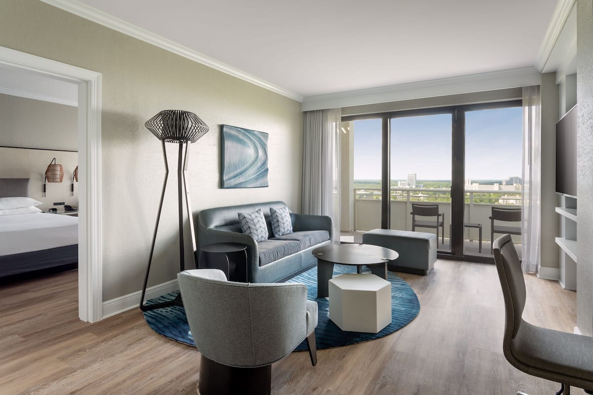 A modern hotel room with a bed, a grey sofa, a coffee table, and a view of the city through large windows.