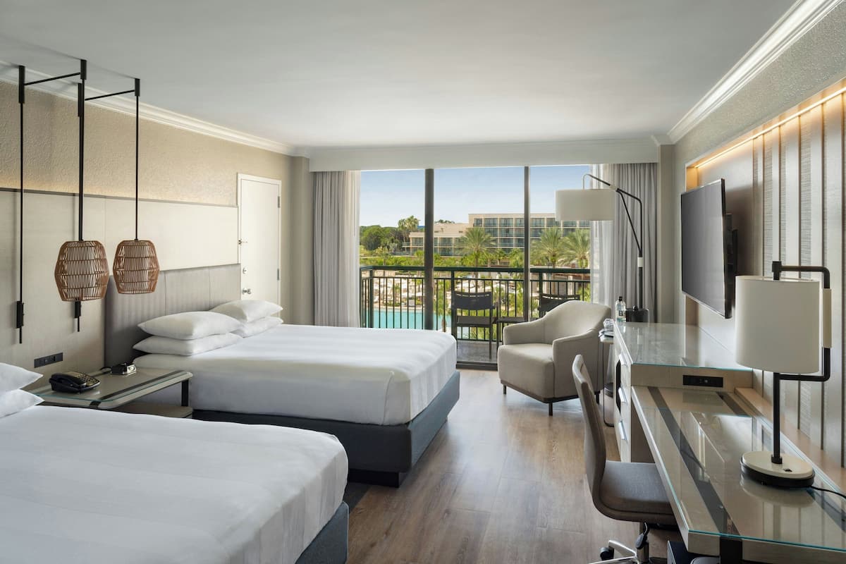 A hotel room with two beds, a desk, a chair, a TV, and a balcony with a view of buildings and greenery.
