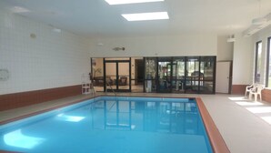 Une piscine intérieure à l’eau claire, dotée d’un sol carrelé et d’une porte vitrée donnant sur un espace extérieur aménagé avec des chaises.