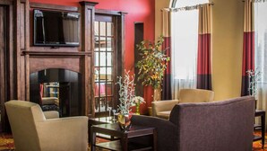 Un hall d’hôtel avec une cheminée, deux fauteuils, un canapé, un téléviseur fixé au mur et une grande fenêtre avec des rideaux.