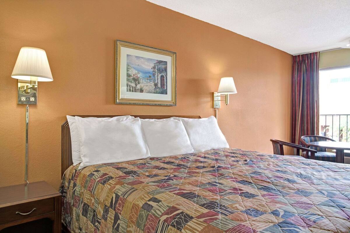 A hotel room with a bed, two bedside lamps, a painting, a desk, and a chair.