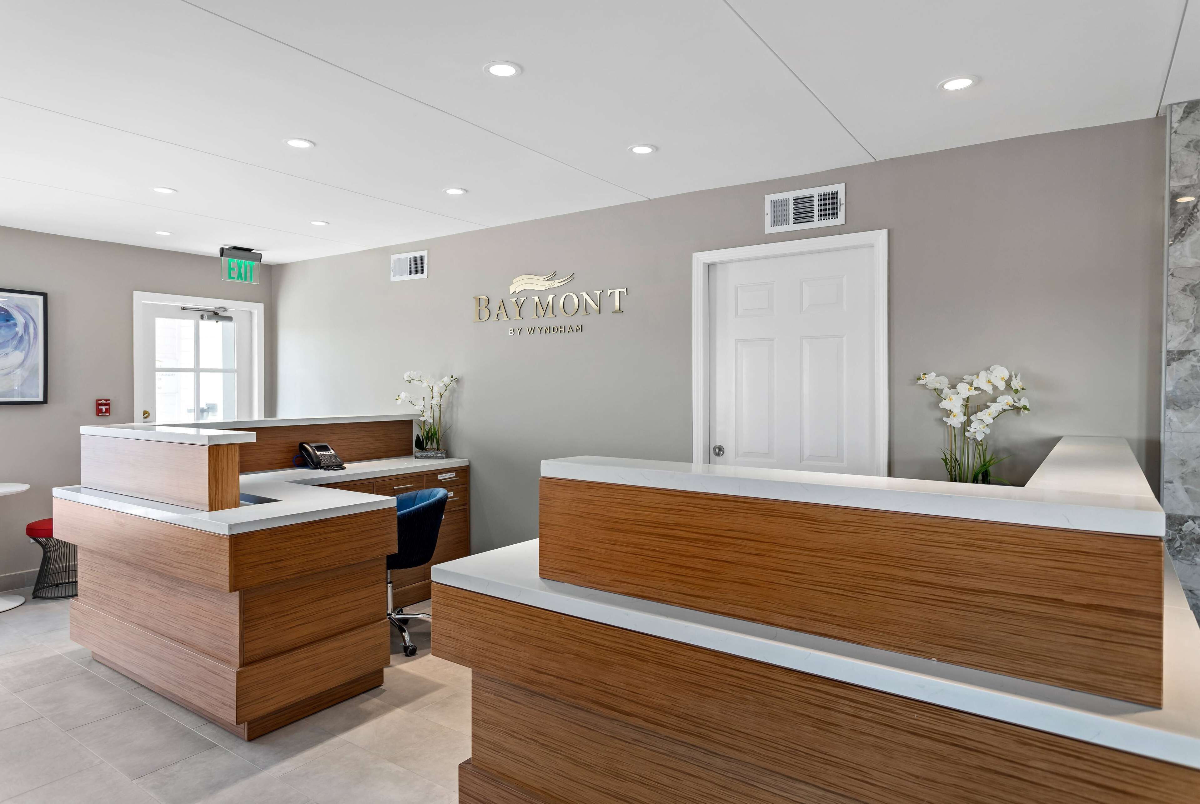Un comptoir d’accueil avec un revêtement en bois, une porte blanche et une enseigne fixée au mur sur laquelle on peut lire « Baymont by Wyndham ».