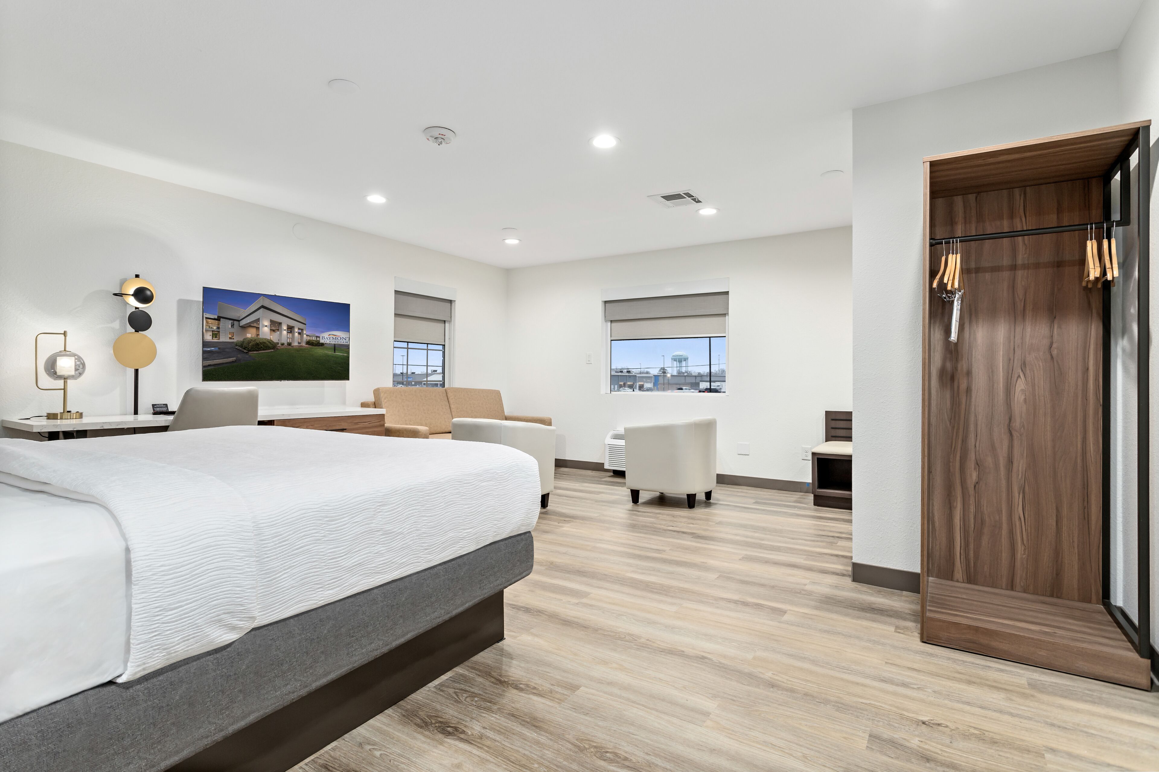 Ein modernes Hotelzimmer mit einem großen Bett, einem Schreibtisch, einem Sessel und Blick auf eine Stadtlandschaft durch große Fenster.