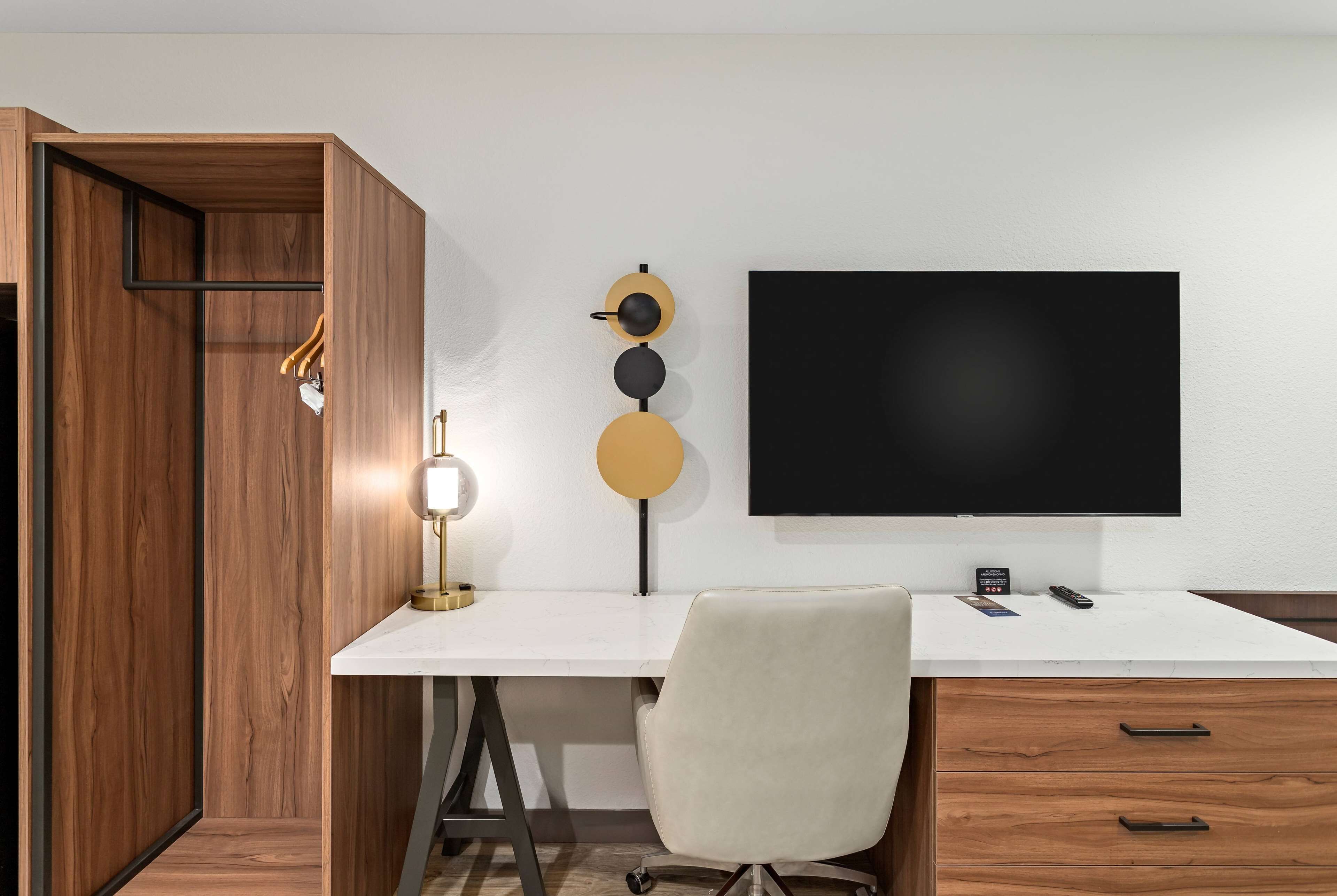Un espacio de oficina moderno con un escritorio blanco, un televisor de pantalla plana grande, un armario de madera y una lámpara de escritorio.