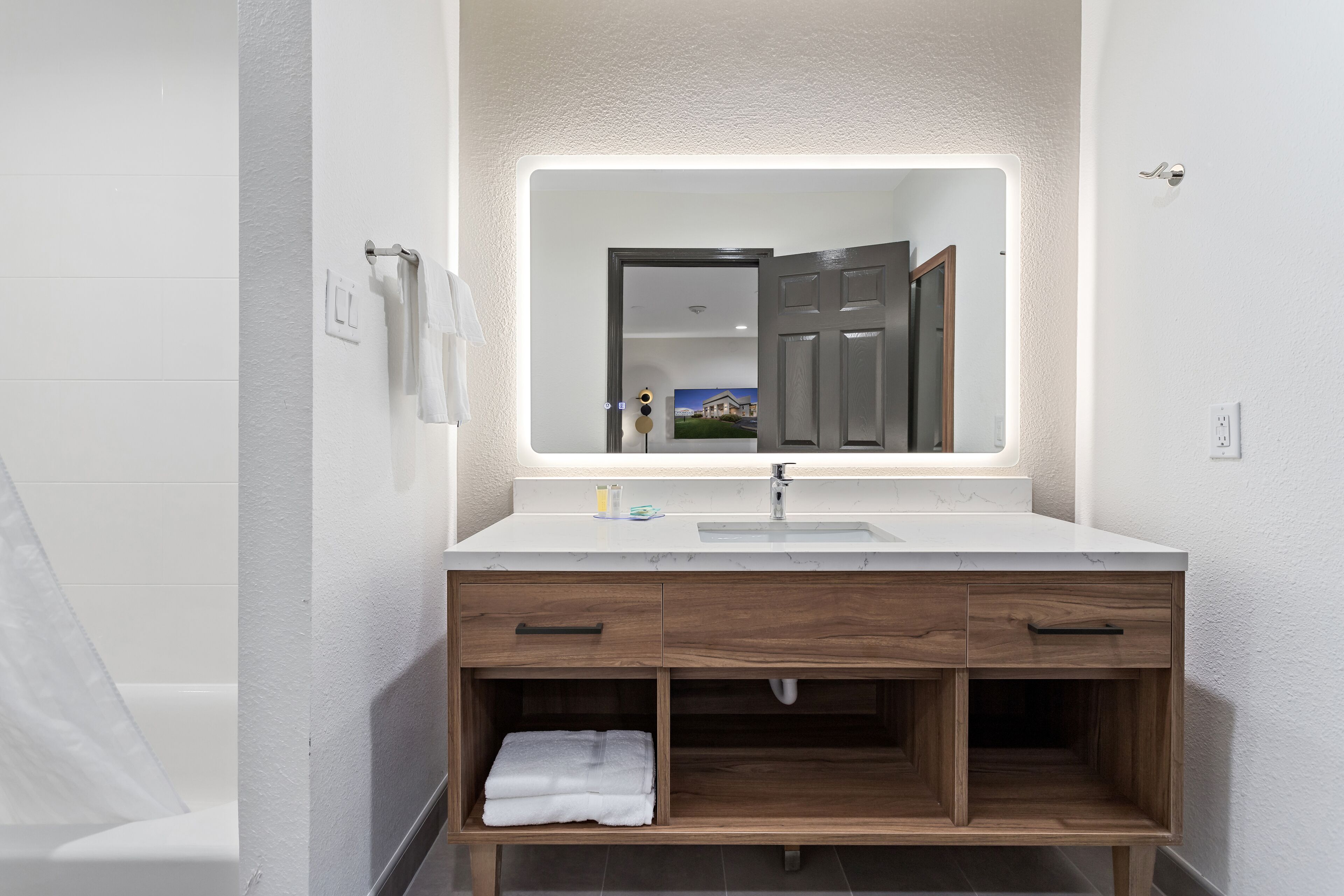A modern bathroom with a white vanity, a mirror with integrated lighting, and a wooden cabinet below.