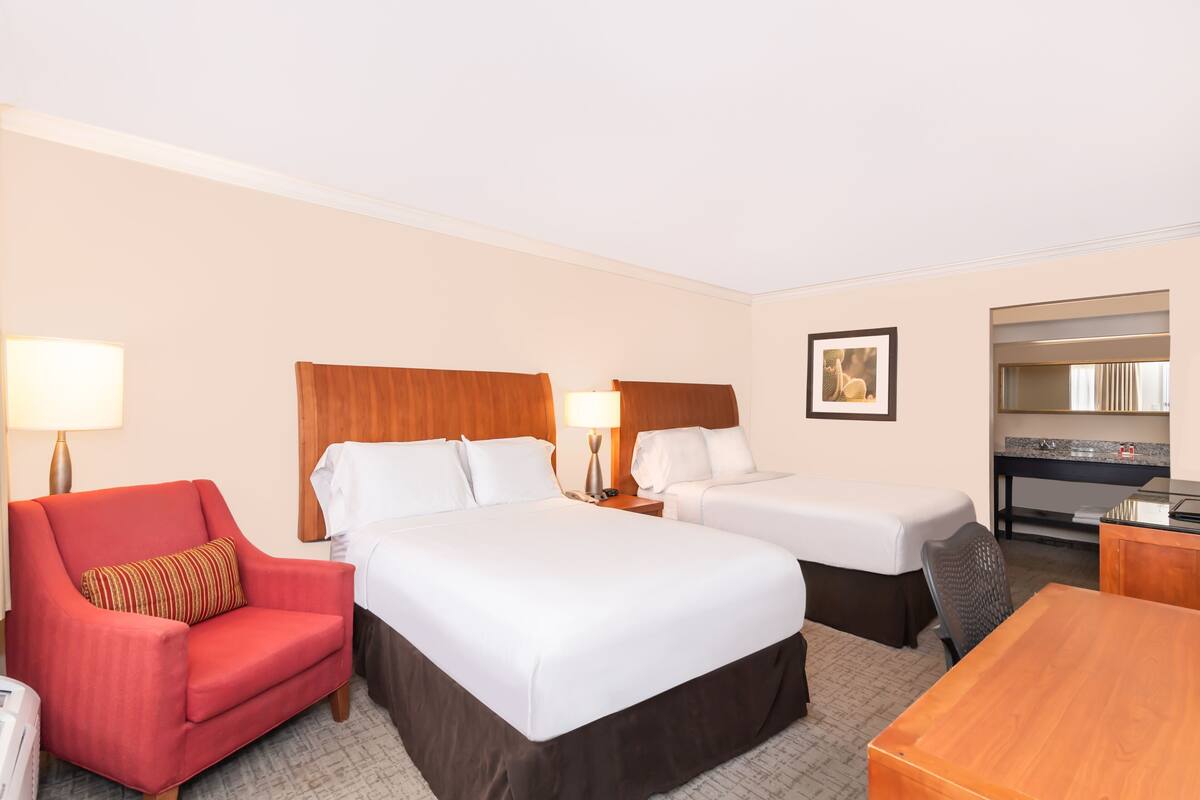 A hotel room with two beds, a red armchair, a wooden desk, and a kitchenette.