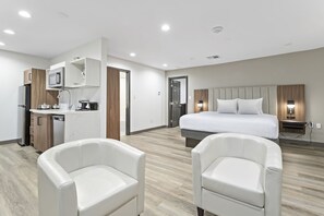 A modern hotel room with a kitchenette, a bed with white bedding, two white leather chairs, and a wooden floor. - Baymont by Wyndham Beaumont (Beaumont)