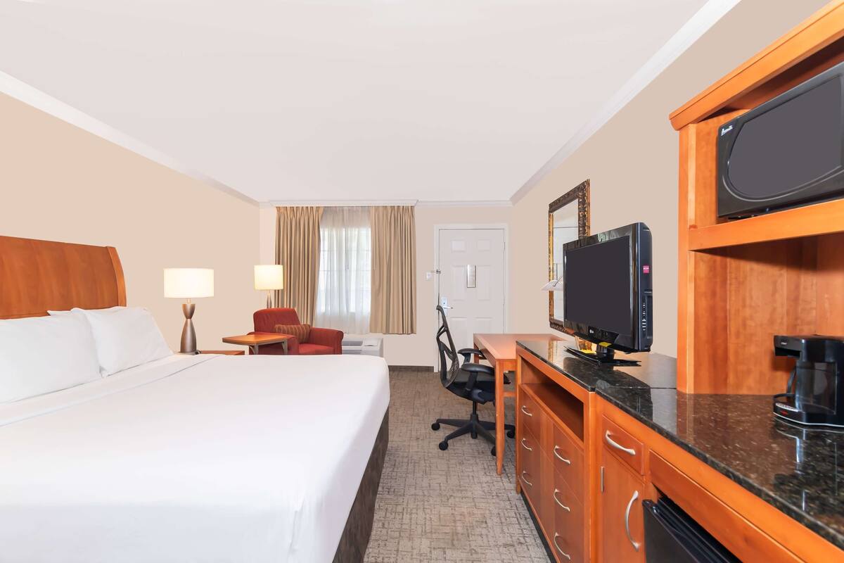 A hotel room with a bed, desk, chair, television, and kitchen area.