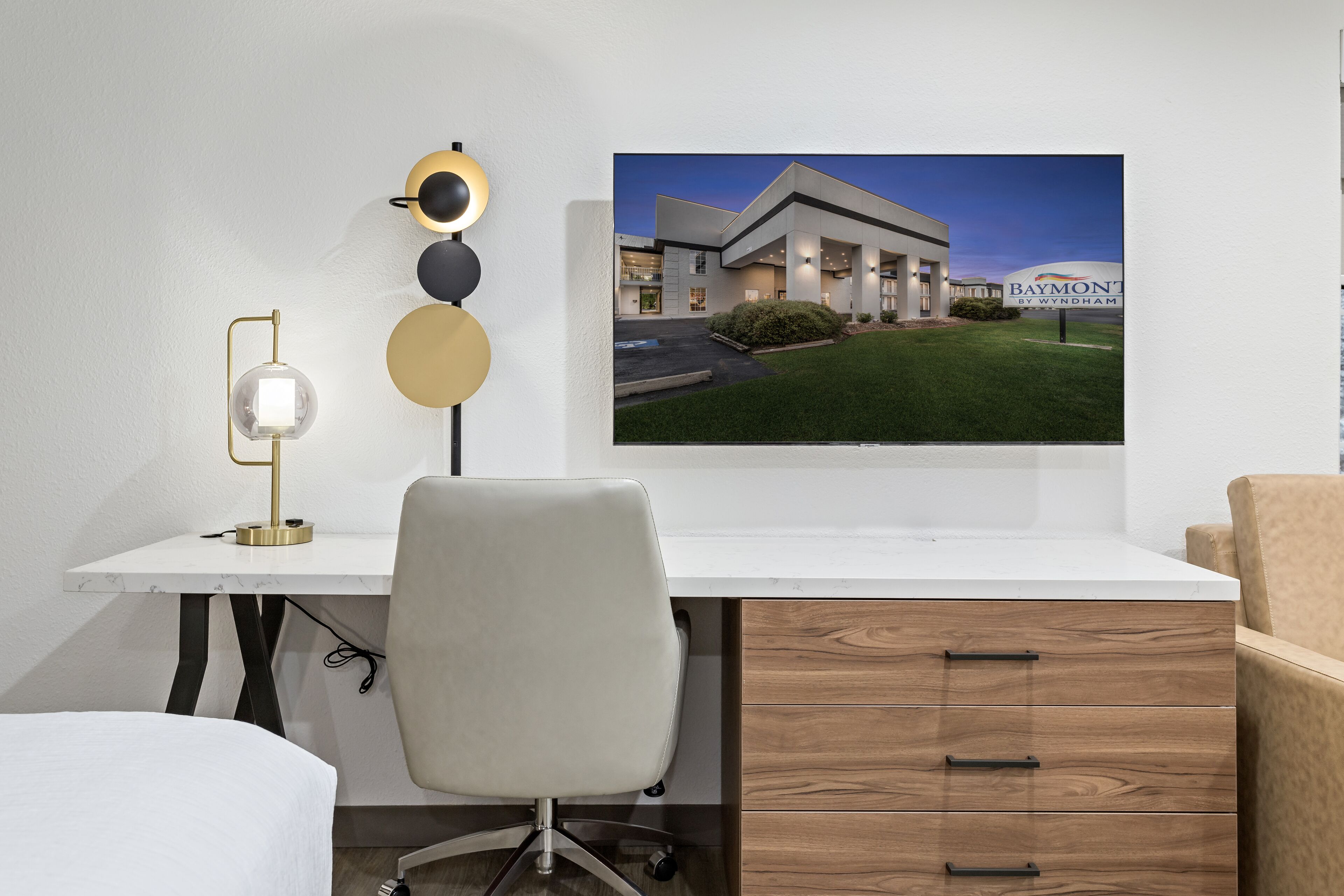 A modern hotel room with a white desk, a chair, a lamp, and a large framed picture of the hotel exterior.