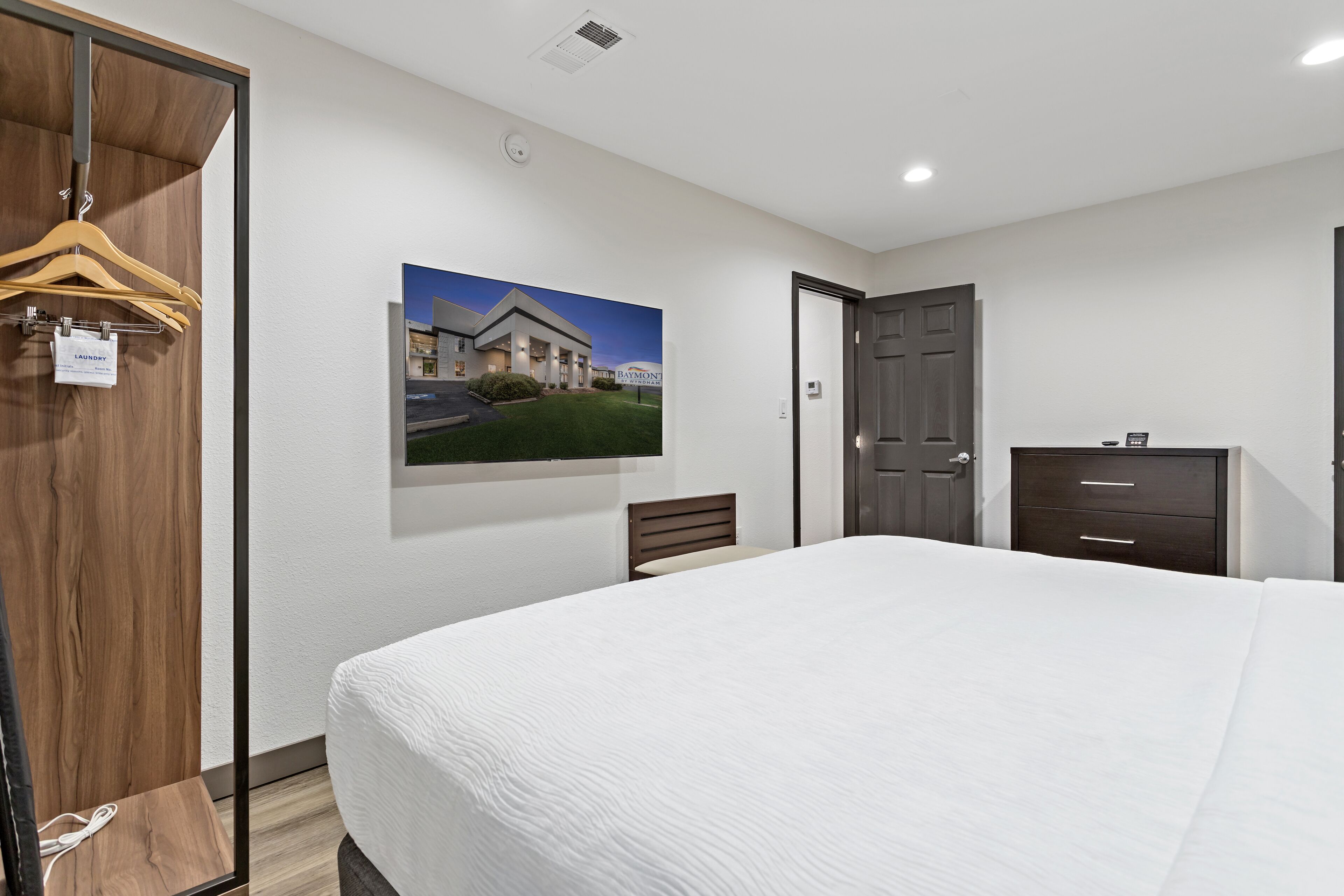 Habitación de hotel con una cama grande, un armario de madera, un escritorio con cajones, una silla y un cuadro en la pared.